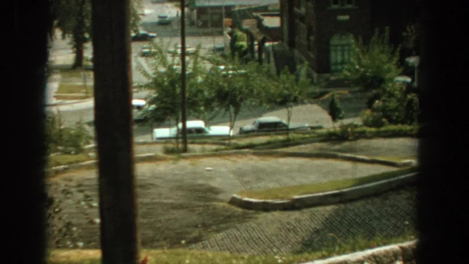 Elevated 1959 Florida street view through vertical window bars, showing parked cars, trees, sidewalks, and low residential buildings.