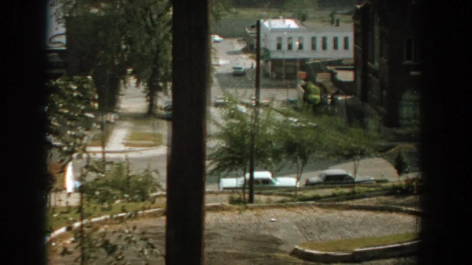 Late-1950s Florida home-movie frame showing cars, trees, a white building, and a curved residential street through dark window bars.
