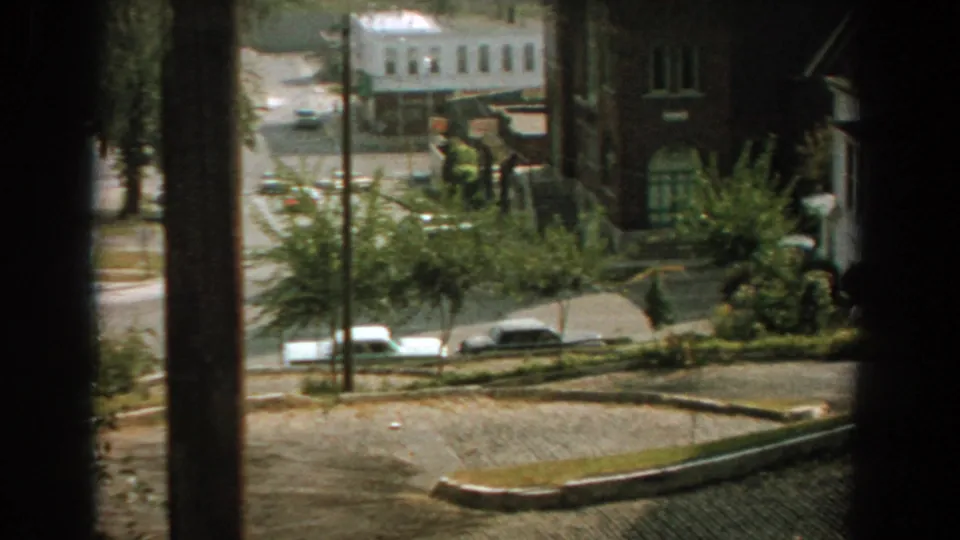 1959 Florida home-movie view across a residential street with parked cars, brick buildings, trees, and foreground posts.