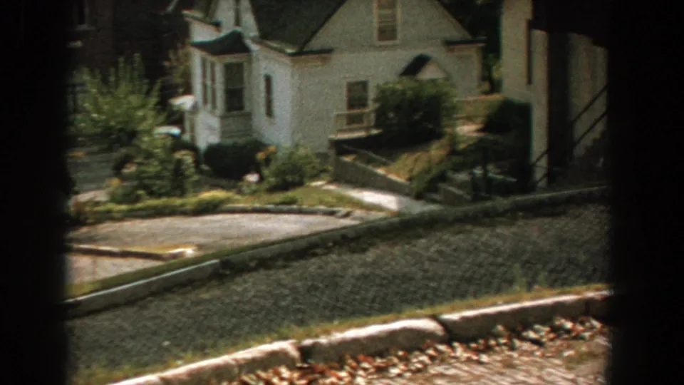 1959 Florida residential view with a white two-story house, paved street, trees, shrubs, and parked cars nearby.