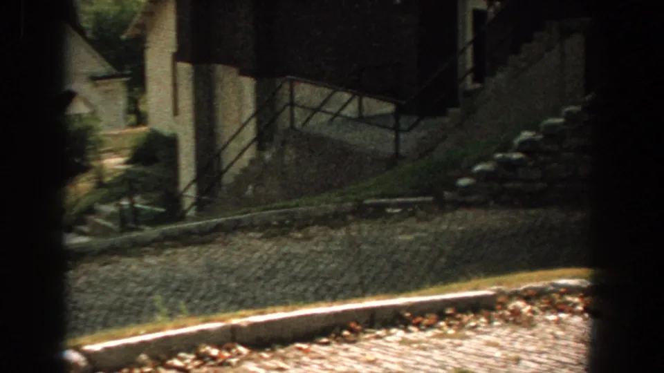 Late-1950s Florida home-movie frame with a street edge, building wall, railing, shrubs, and shadowed foreground obstruction.