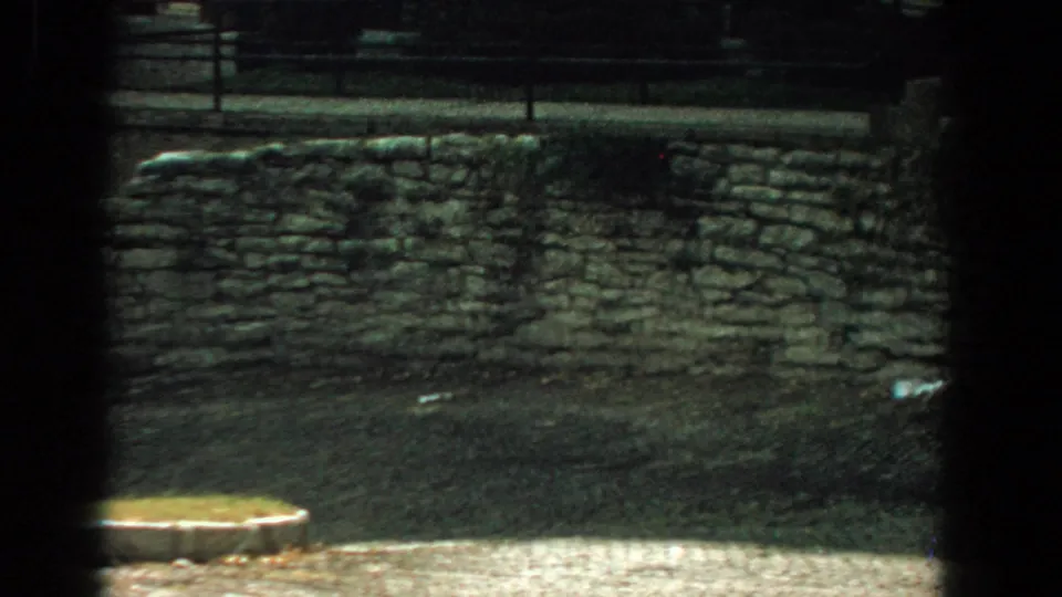1959 Florida 8mm frame showing a long low stone wall, paved foreground, grass, railing, and dark borders suggesting a shaded viewpoint.