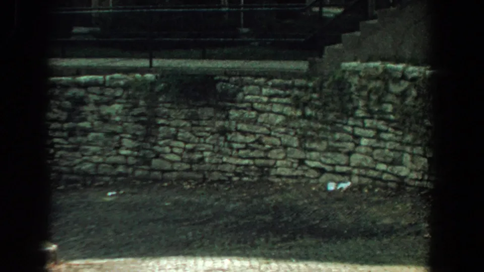1959 Florida 8mm frame of a low stone wall, shadowed ground, stairs, and railings, filmed from an obstructed exterior viewpoint.
