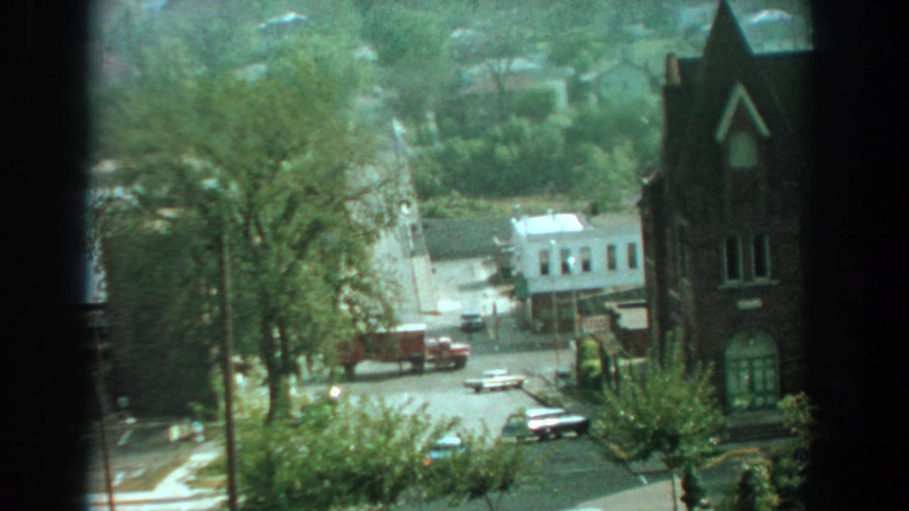 Elevated view of a sunny street intersection with mid-century cars, trees, and a brick church-like building.