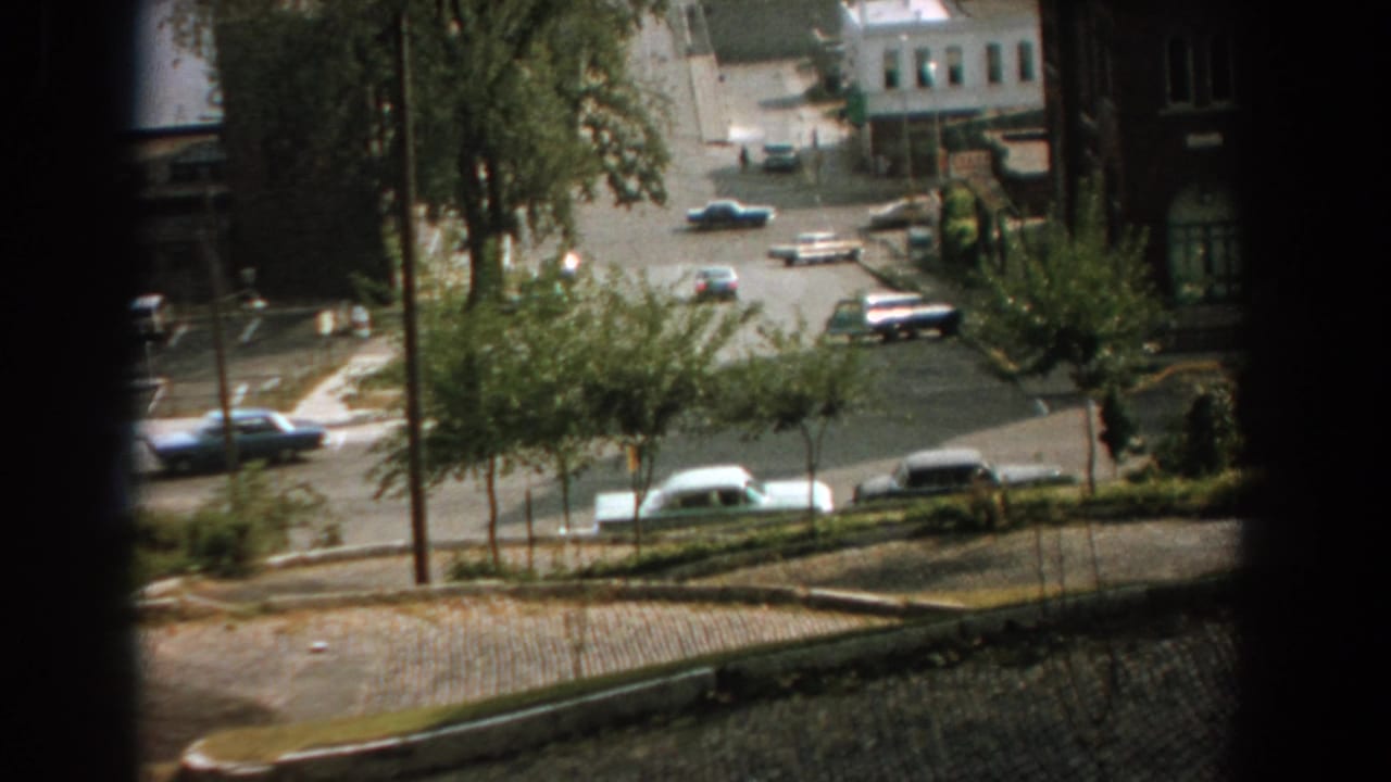 High-angle view of a quiet city intersection with several cars, trees, sidewalks, and dark window framing.