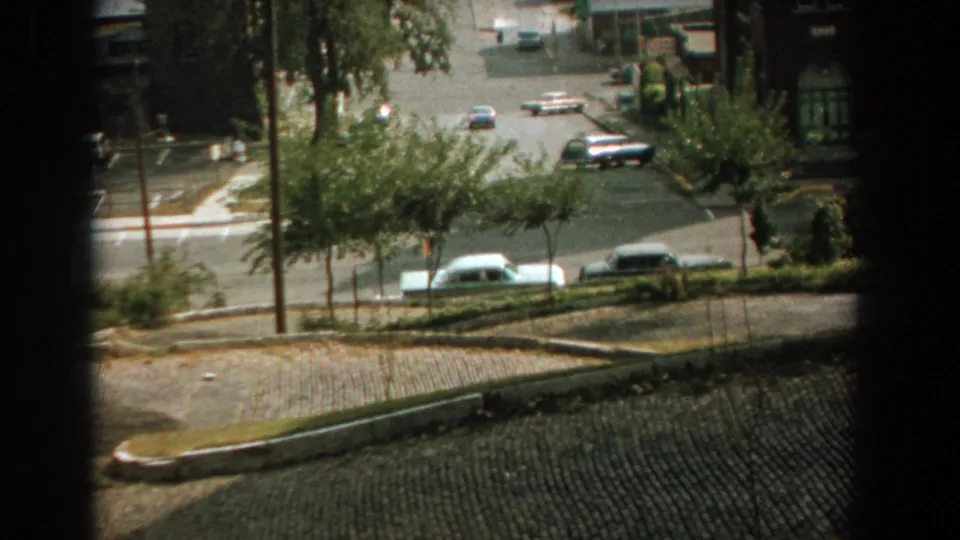 High-angle 1959 Florida street view with a white sedan, darker cars, trees, and sidewalks near a quiet intersection.