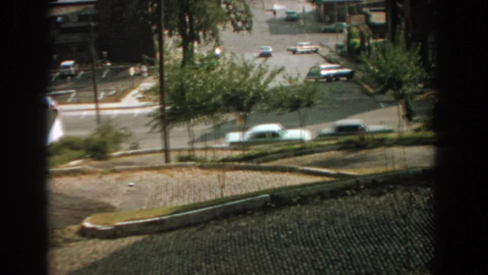 1959 Florida home-movie frame showing sparse city traffic through a dark window opening, with sidewalks and trees along the road.