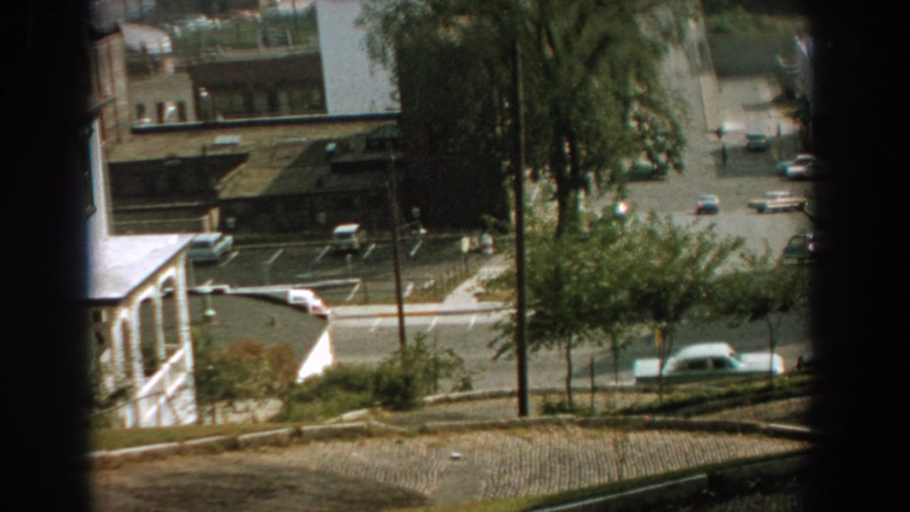 High-angle street view with cars, trees, buildings, and a dark window edge.