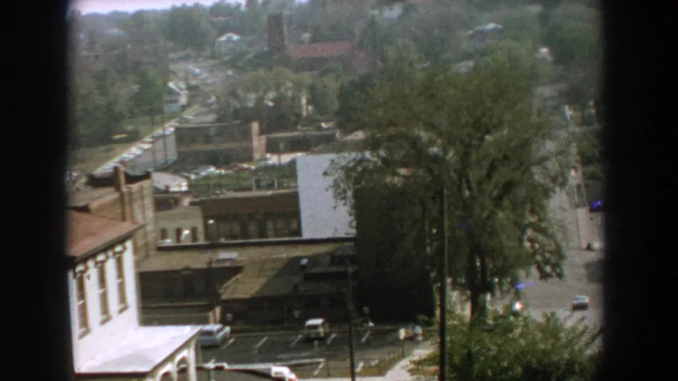 1959 Florida home-movie frame with an elevated look over rooftops, trees, a small parking area, and cars along the road.