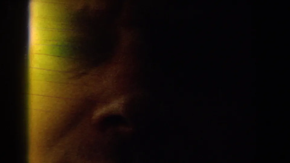 Underexposed 1959 8mm frame with a partial face or reflection, warm flare, and dark interior-like shadow.