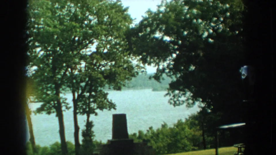 A Florida river is seen through leafy trees, with a small stone monument or marker near the grassy foreground in the 1959 home-movie frame.