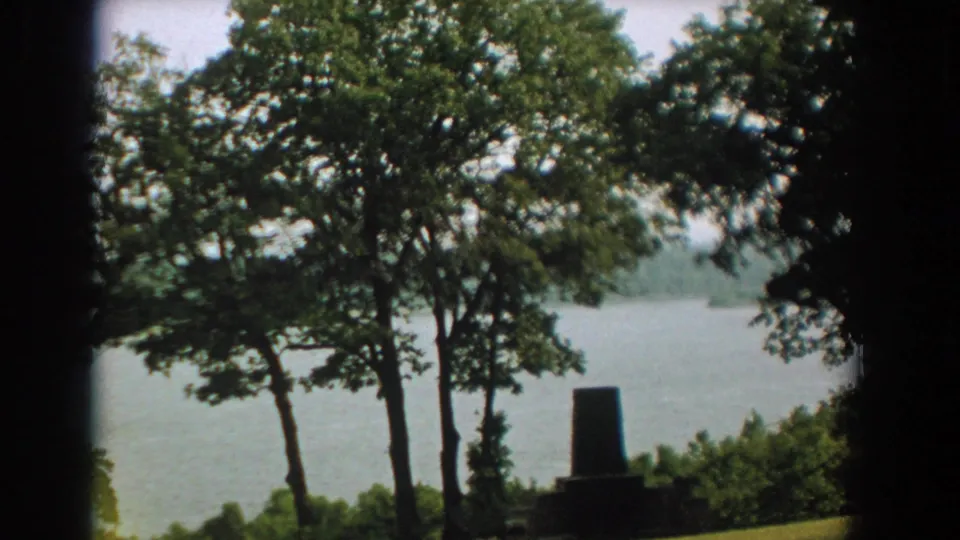 A soft 1959 8mm frame shows trees in front of a Florida river, with a small stone marker or monument near the lower right foreground.