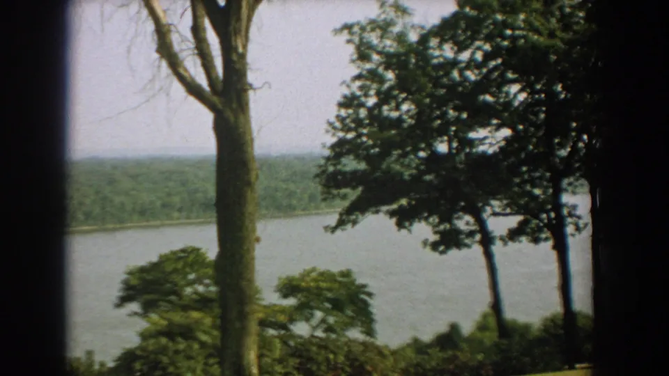 1959 Florida home-movie frame with a broad waterway, dense green far shore, and trees silhouetted at the edges.