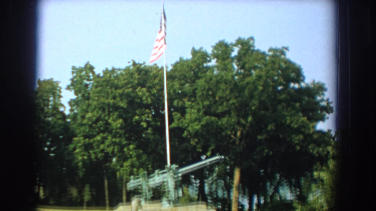 1959 Florida 8mm frame showing a U.S. flag above a mounted artillery gun in a grassy outdoor setting with dense trees.