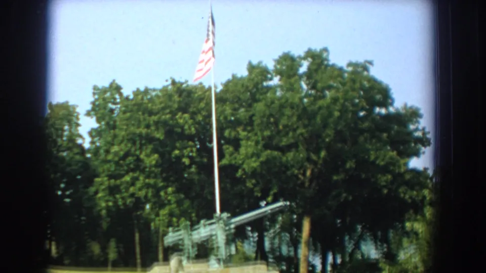 1959 Florida 8mm frame showing a U.S. flag above a mounted artillery gun in a grassy outdoor setting with dense trees.