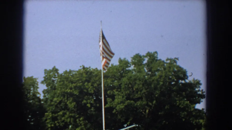 1959 Florida home-movie frame with the U.S. flag centered above green trees and a partial artillery display at the lower edge.