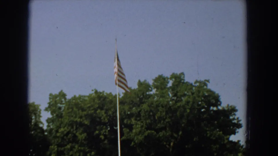 1959 Florida 8mm frame showing the U.S. flag on a tall pole against trees and sky, with the monument mostly out of view.