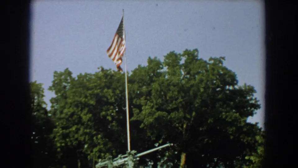 1959 Florida frame of a U.S. flag above a mounted artillery gun, with leafy trees filling the background.