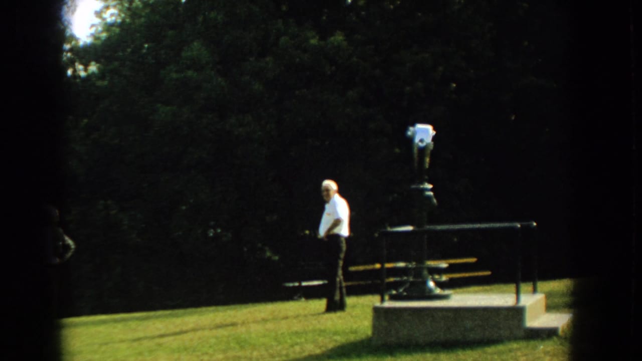 In 1959 Florida home-movie footage, an older man in a white shirt stands by a coin-operated viewer and bench in a tree-lined park.