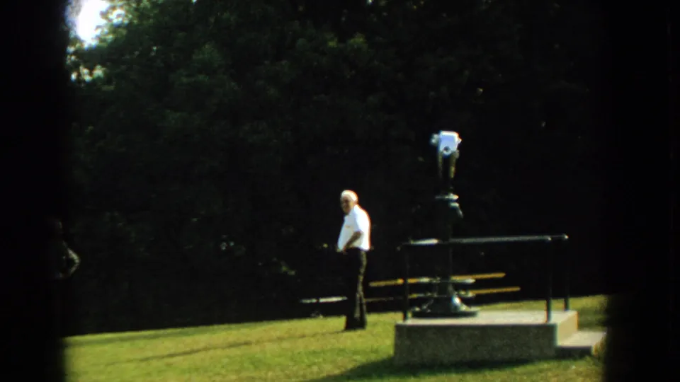 In 1959 Florida home-movie footage, an older man in a white shirt stands by a coin-operated viewer and bench in a tree-lined park.