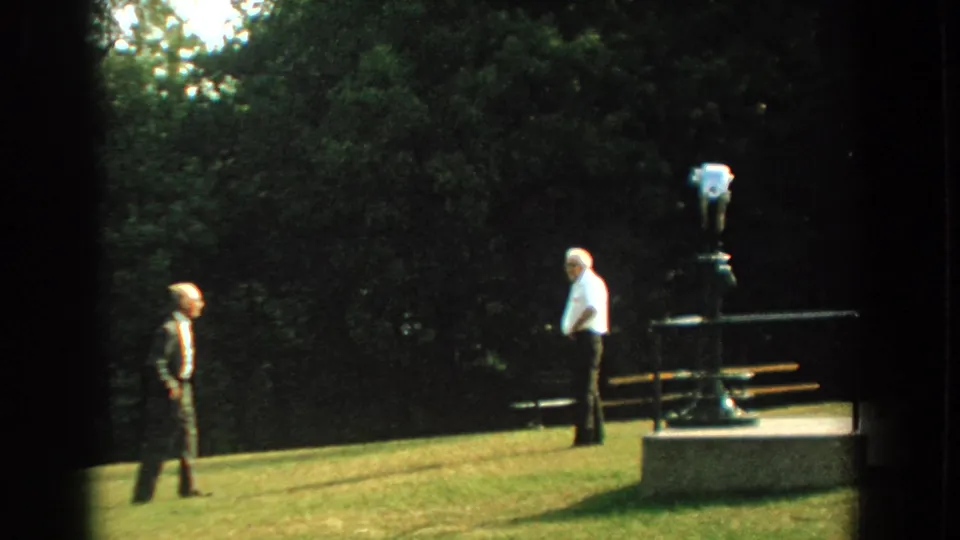 Two older men appear in conversation on a grassy Florida park slope in 1959, near a coin-operated viewer and bench.