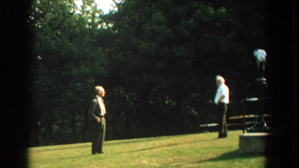 A suited older man stands on the grassy slope as another older man remains near the park viewer in this 1959 Florida 8mm frame.