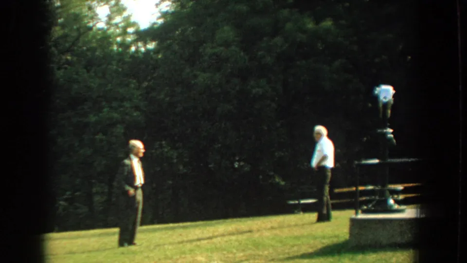 Two older men face each other beside a raised coin-operated viewer platform in a tree-lined Florida park, recorded on 8mm film in 1959.