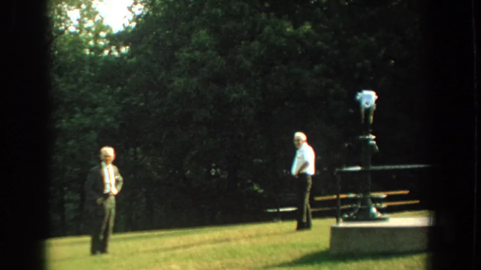 In 1959 Florida home-movie footage, two adult men stand on a grassy park slope near a mounted binocular viewer.