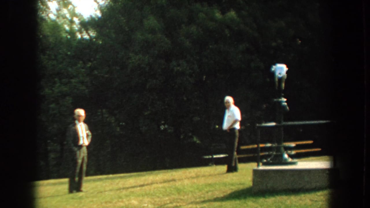Two men stand on a lawn with a mounted viewer at right and trees behind.