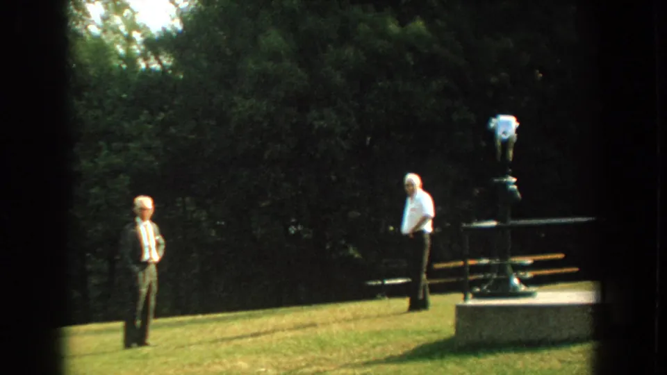 Two adult men remain separated on a Florida park lawn, with a coin-operated binocular viewer on a concrete base at right.