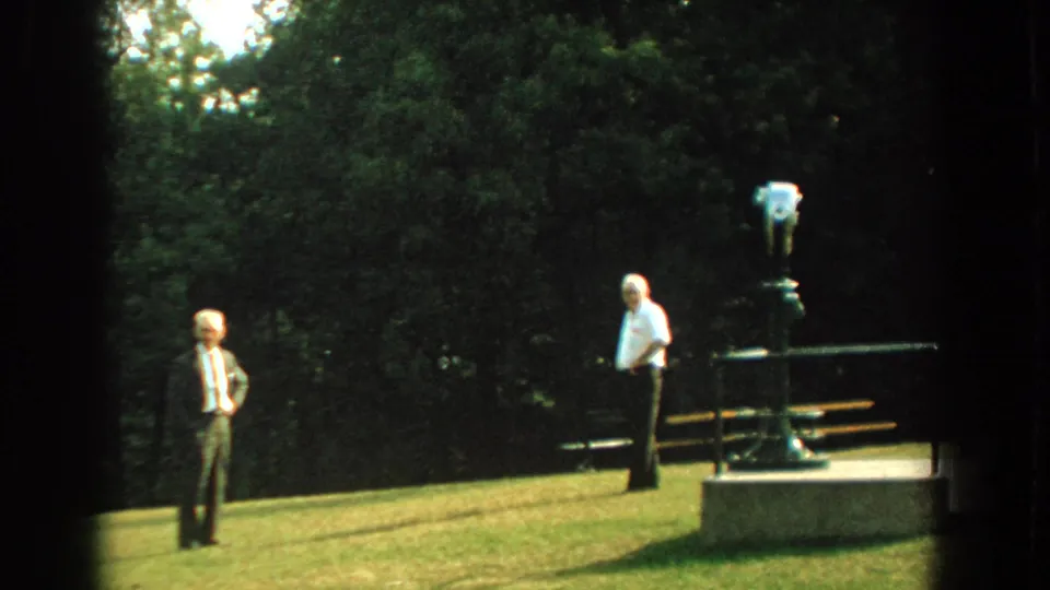 The 8mm frame shows two men standing apart on a sunny grassy hillside beside a public binocular viewer.
