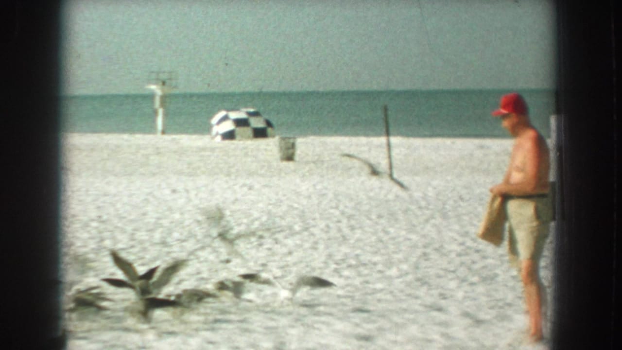 1959 Florida beach frame with a man in a red cap watching seagulls near checkered umbrellas and the ocean horizon.