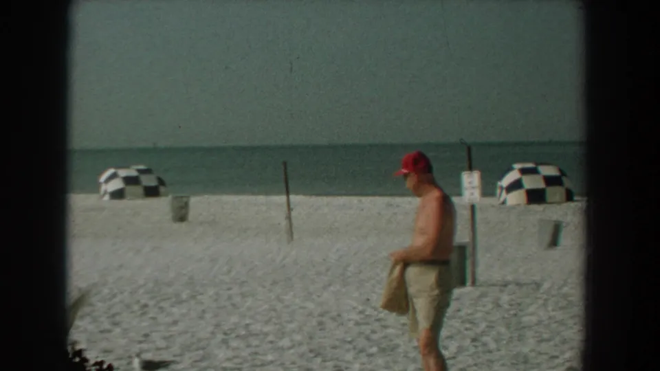 1959 Florida home-movie frame showing a man in a red cap and shorts crossing the sand near checkered beach umbrellas.