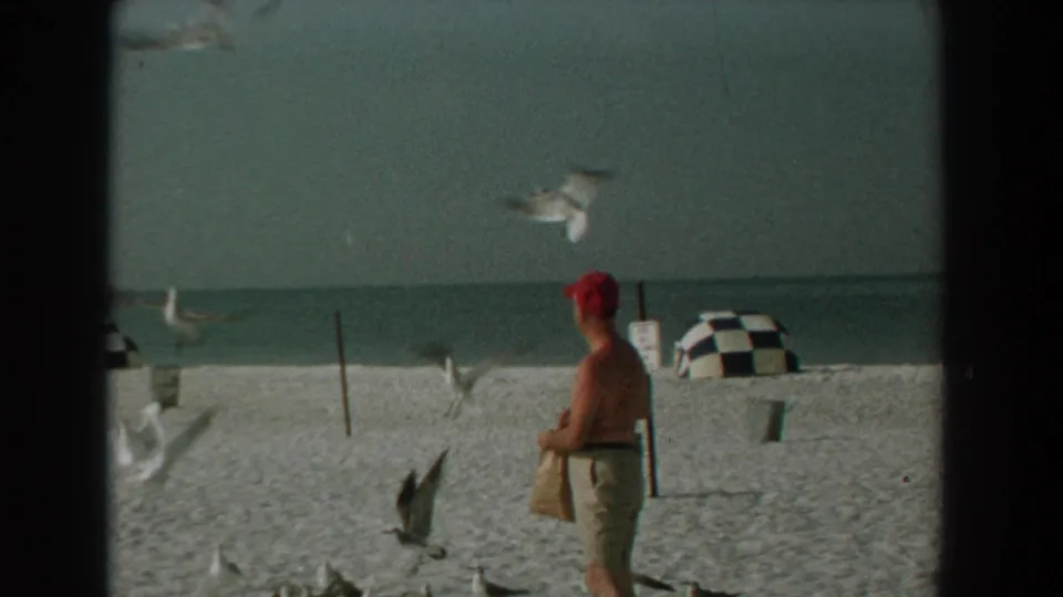 1959 Florida beach frame with gulls in flight, a man in a red cap, checkered umbrellas, and the ocean beyond.