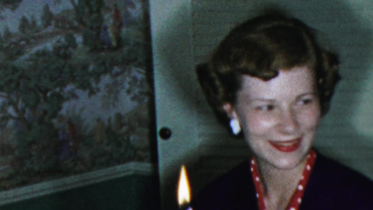 In 1959 Paramus home-movie footage, a woman with curled hair, white earrings, and red accessories smiles near a lit candle indoors.