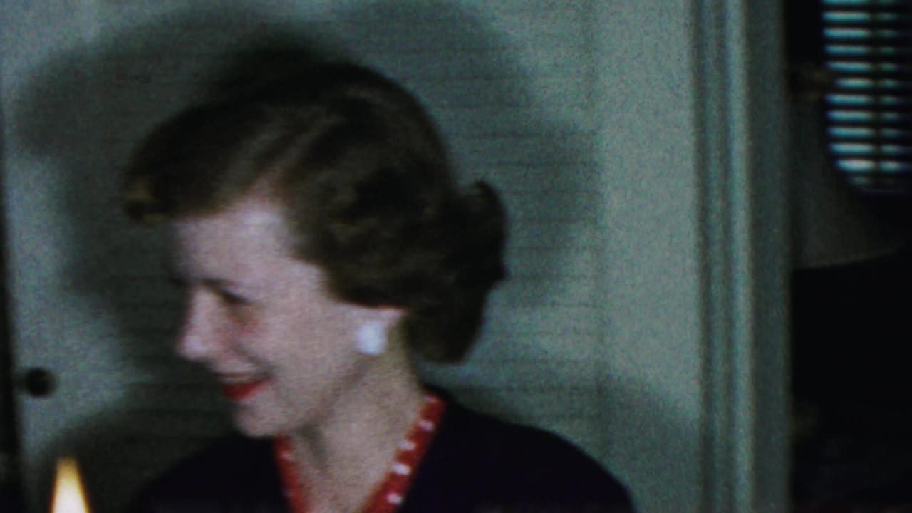 Two adult women in 1959 Paramus home-movie footage smile toward each other near candlelight in a private interior.