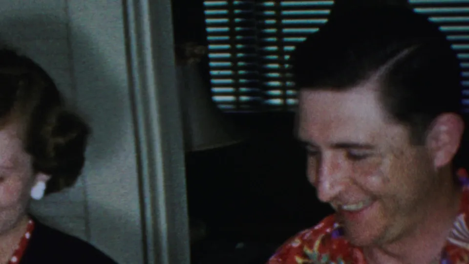 An adult man in a colorful floral shirt laughs in a private Paramus interior, with a woman partly visible at the edge of the frame.