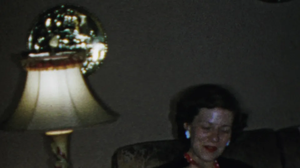 Adult woman smiles on upholstered seating next to a lit lamp in a 1959 Paramus, New Jersey home interior.