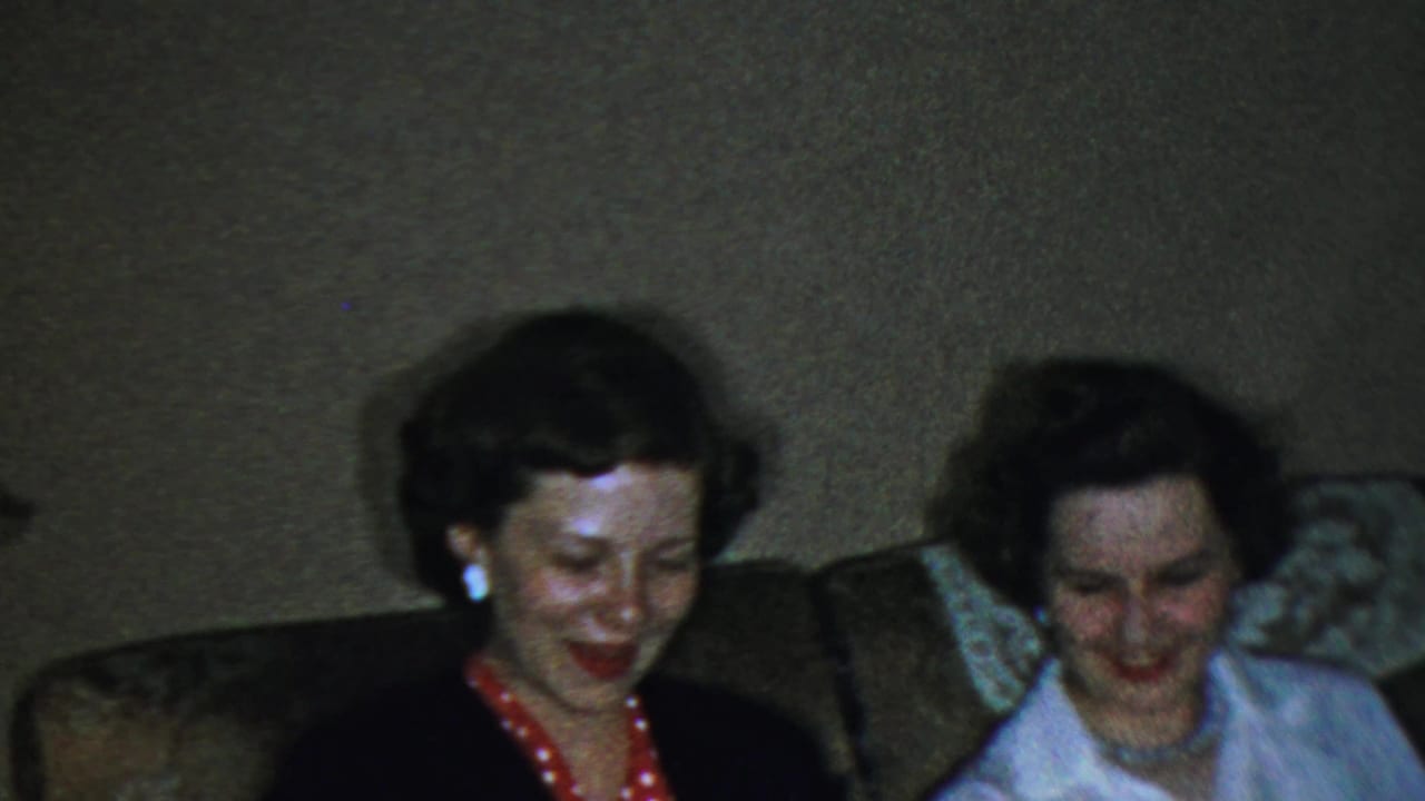 Two adult women sit close together on a couch in a 1959 Paramus home movie, smiling during a relaxed indoor moment.