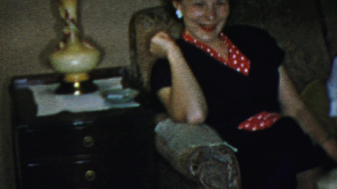 Adult woman in a dark dress with red polka-dot accents sits in an armchair beside a side table and lamp in a 1959 Paramus living room.