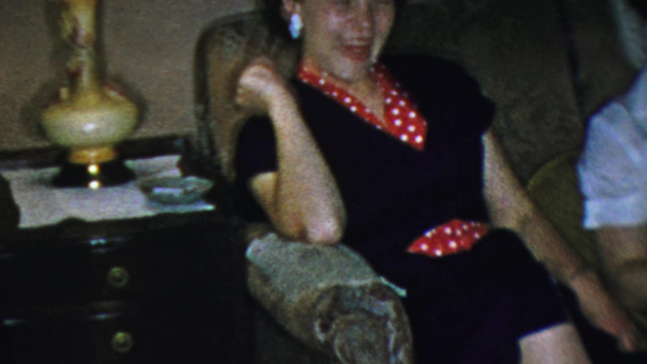 In a 1959 Paramus living room, a woman in a dark dress with red polka-dot trim laughs from a sofa near a side table and lamp.