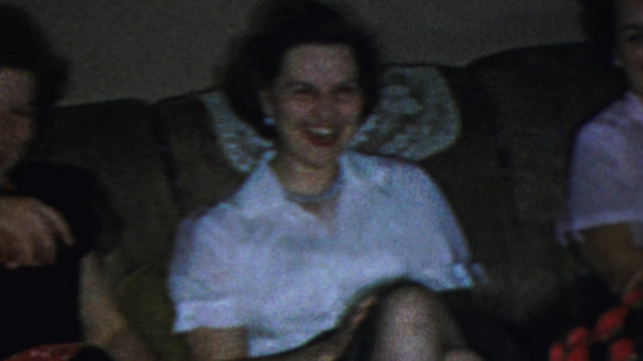 A smiling woman in a white blouse sits on a living room sofa in the 1959 Paramus home-movie scene, partly framed by other adults.