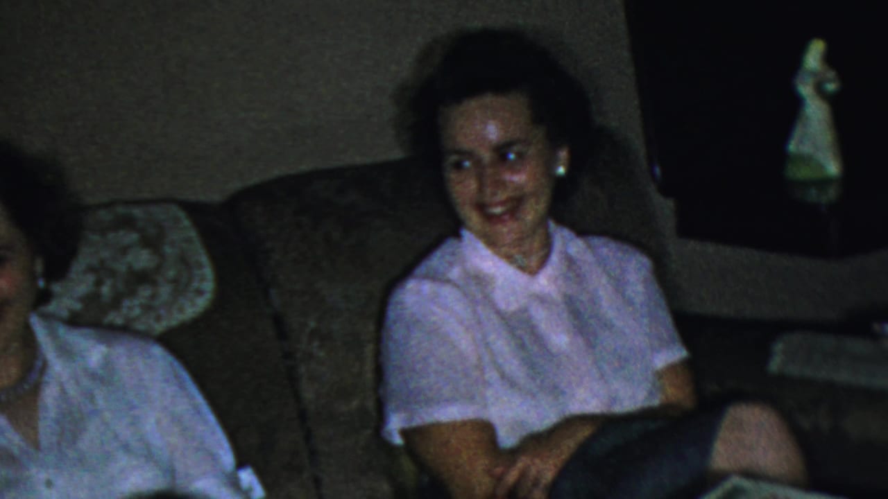 A woman in a white blouse smiles from a sofa in the 1959 Paramus living room as another person leans partly into the frame.