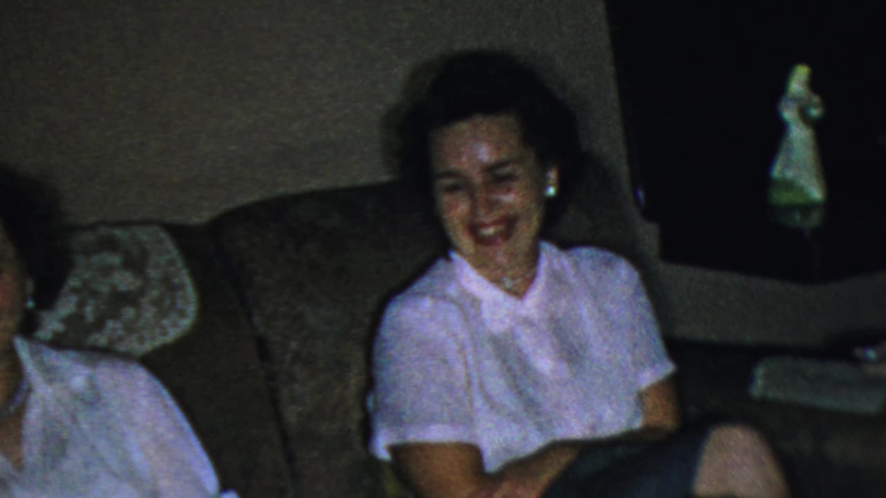Two adult women sit on a brown couch in a dim living room, one smiling toward the camera.