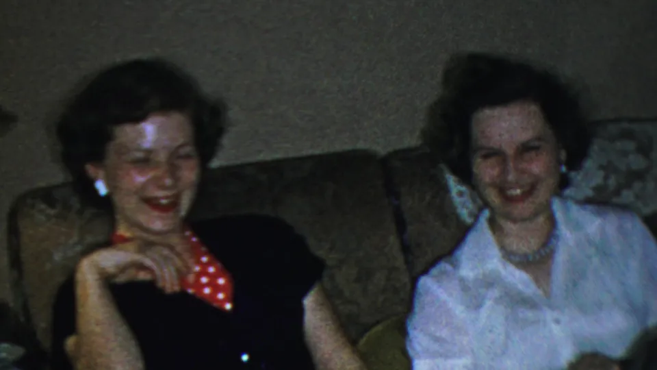 Three adult women laugh together on a couch in the 1959 Paramus home-movie scene, including one wearing a dark top with red polka-dot detail.