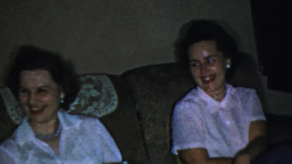 Two adult women smile and laugh on a brown couch in 1959 Paramus home-movie footage, with lace covers visible behind them.