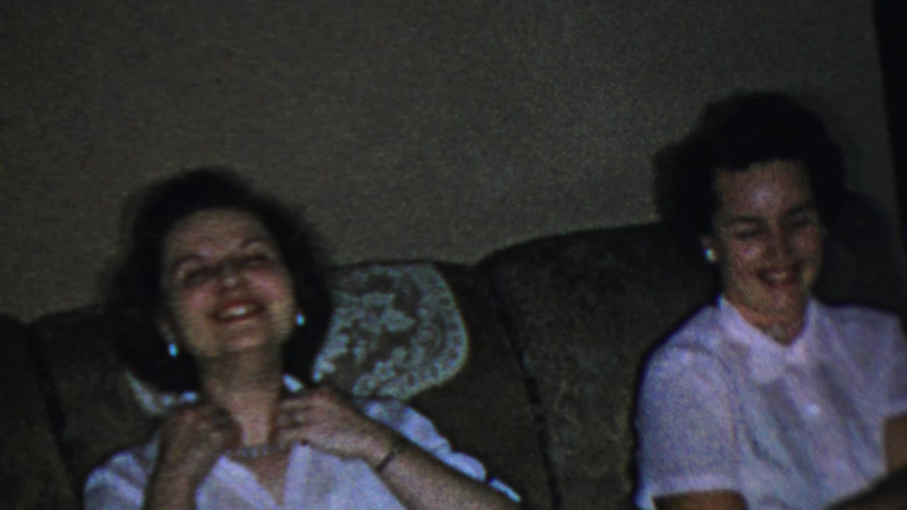 Adult woman in a white blouse laughing on a brown sofa in a dim living room.