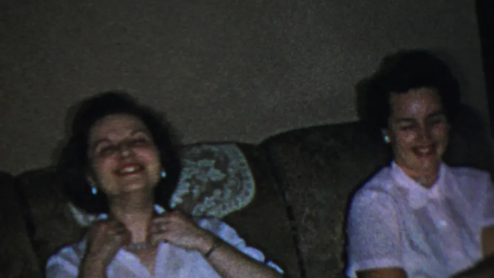 In 1959 Paramus home-movie footage, an adult woman laughs on a brown sofa while wearing a white blouse and necklace.