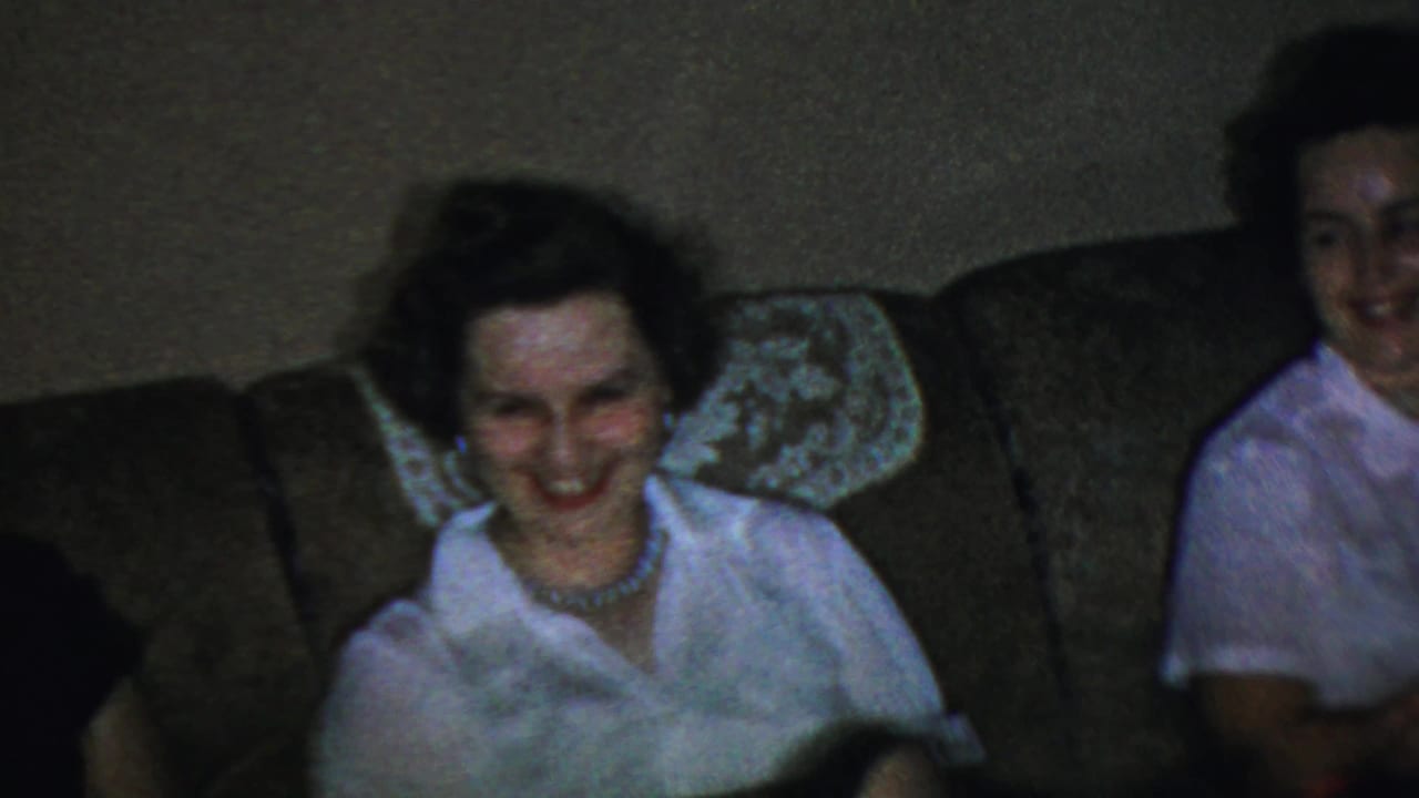 The closing frame shows two adult women on a brown sofa in the supported Paramus 1959 home-movie interior, one smiling downward.