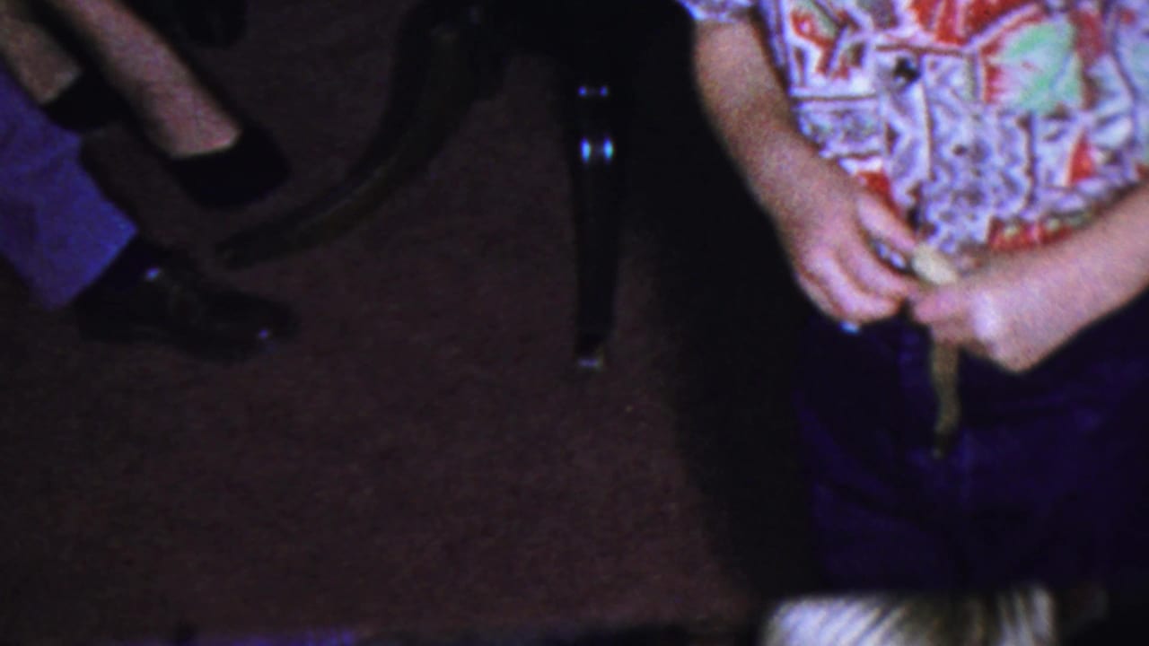 Person in blue pants and patterned shirt stands on brown carpet with hands near a small item.